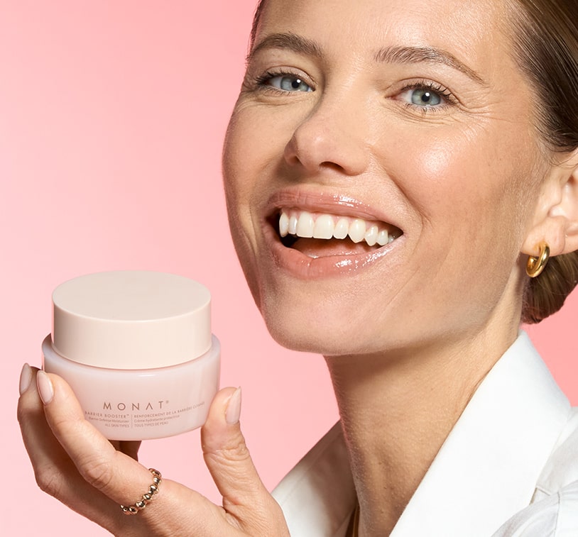 Smiling woman holding MONAT Barrier Booster™ Barrier Defense Moisturizer jar near her cheek.