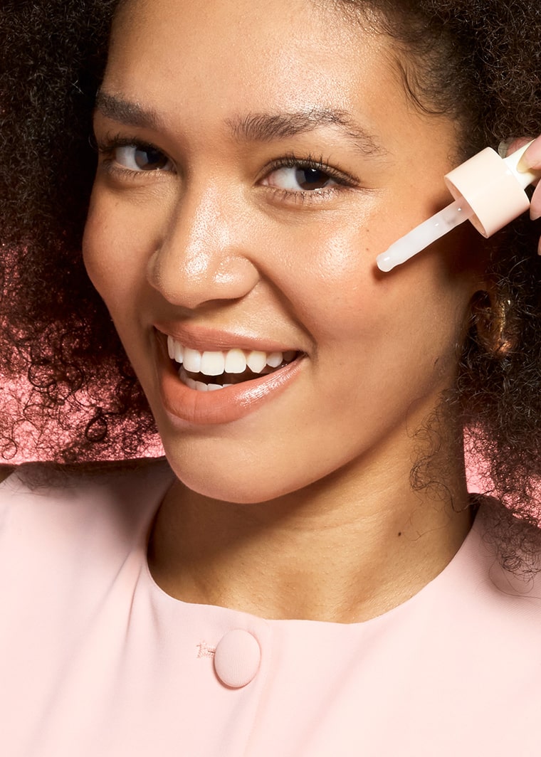 Smiling woman with curly hair holding MONAT Barrier Booster™ Niacinamide Glow Serum dropper bottle near her face.