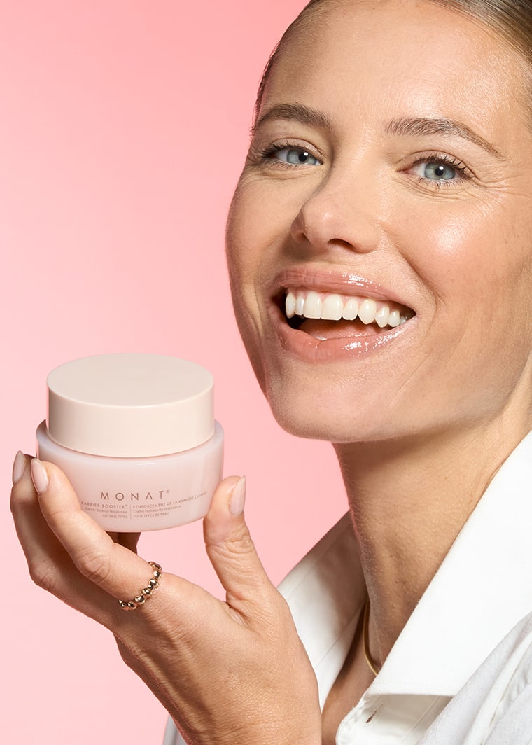 Smiling woman with pulled-back hair applying MONAT Barrier Booster™ Barrier Defense Moisturizer to her cheek.