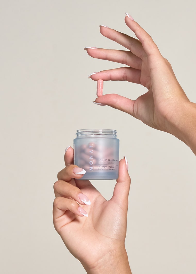 Hands holding pink supplement capsule above open MONAT IR HAIR SUPPLEMENT