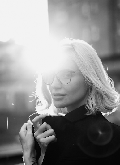 An artistic black and white photograph of a person with short blonde hair wearing glasses and a dark coat