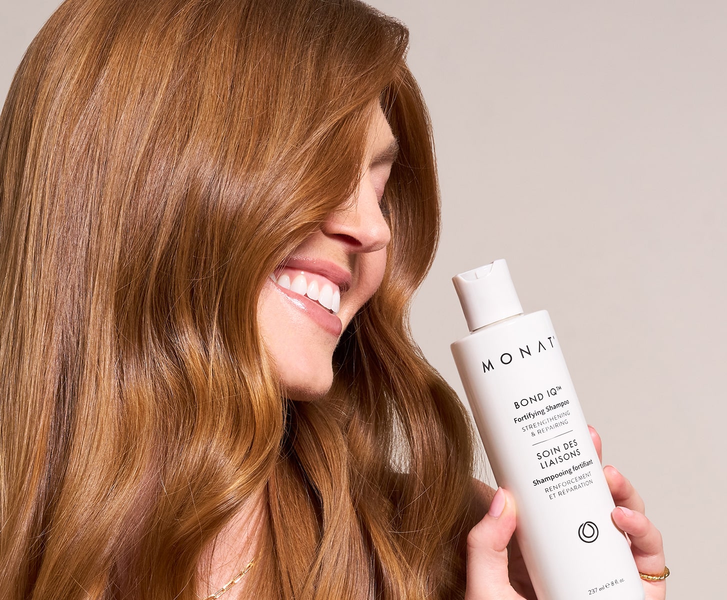 A woman with long, glossy brown hair smiles while holding a white bottle of MONAT Bond IQ Fortifying Shampoo.