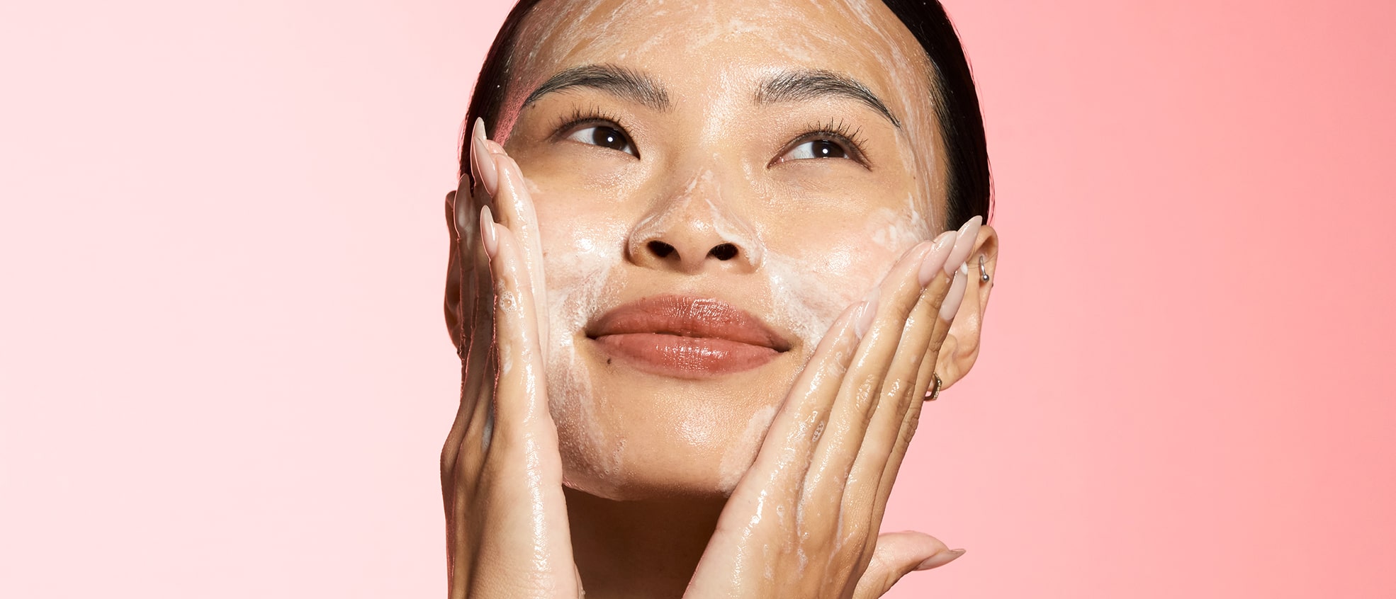 Asian woman with radiant skin applying cleanser to her face with both hands, smiling with eyes closed against pink background