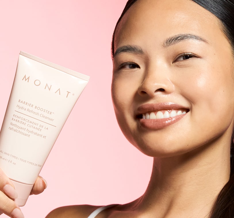 Smiling model with glowing skin holding A MONAT Barrier Booster Refresh Cleanser tube 
