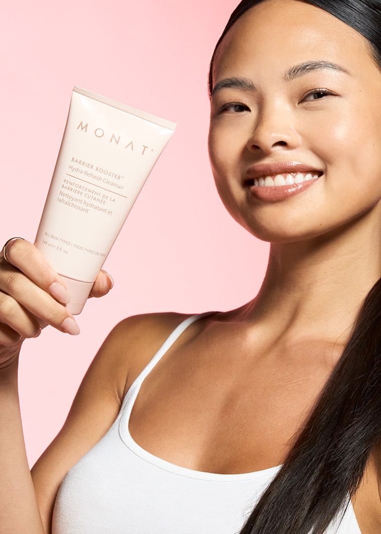 Smiling Asian woman with radiant, glowing skin holding MONAT Barrier Booster Hydra Refresh Cleanser tube, showcasing healthy complexion against soft pink background