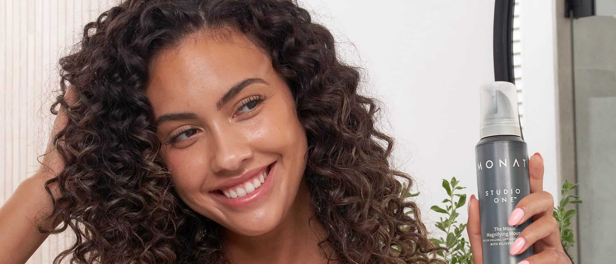 Brunette female with curls holding MONAT STUDIO ONE™ The Moxie™ Magnifying Mousse.