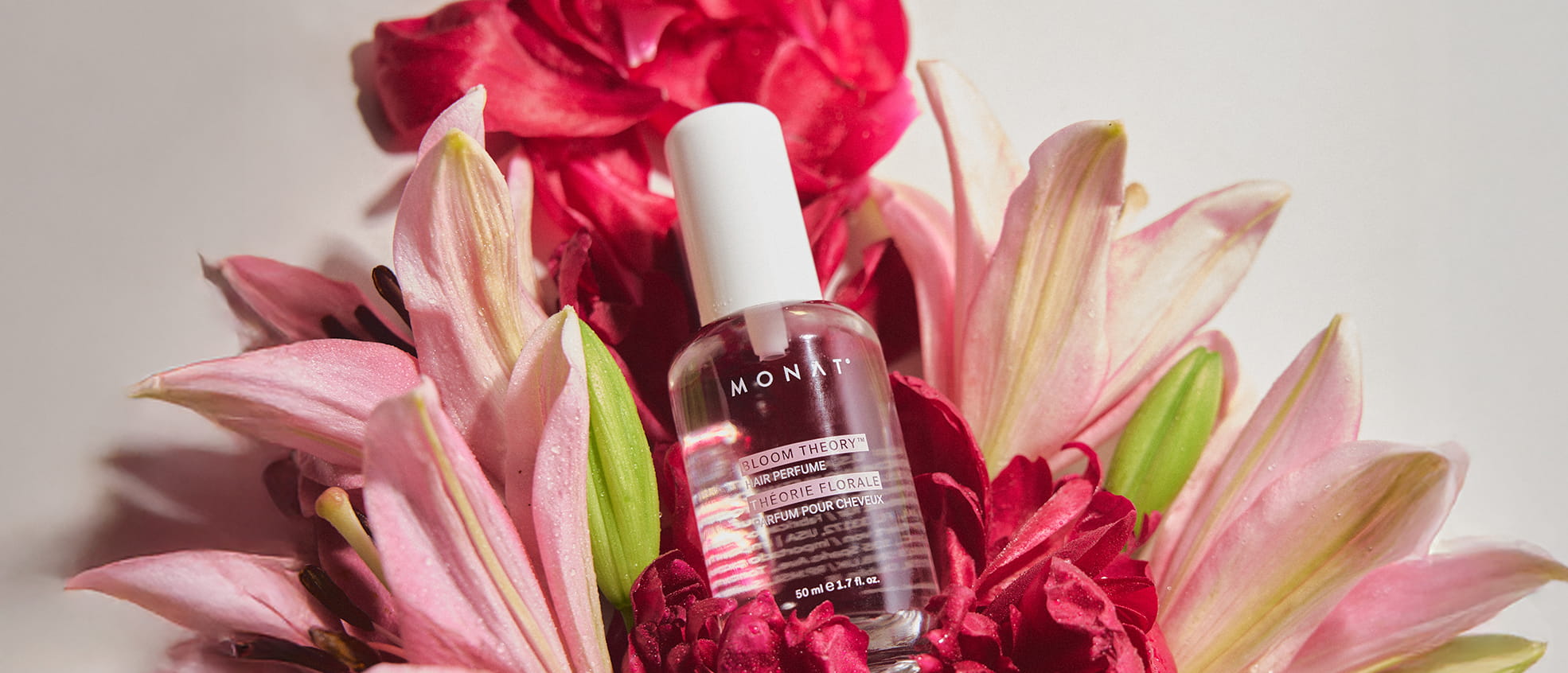 MONAT Bloom Theory Hair Perfume spray bottle nestled among pink lilies and red peonies against a light background