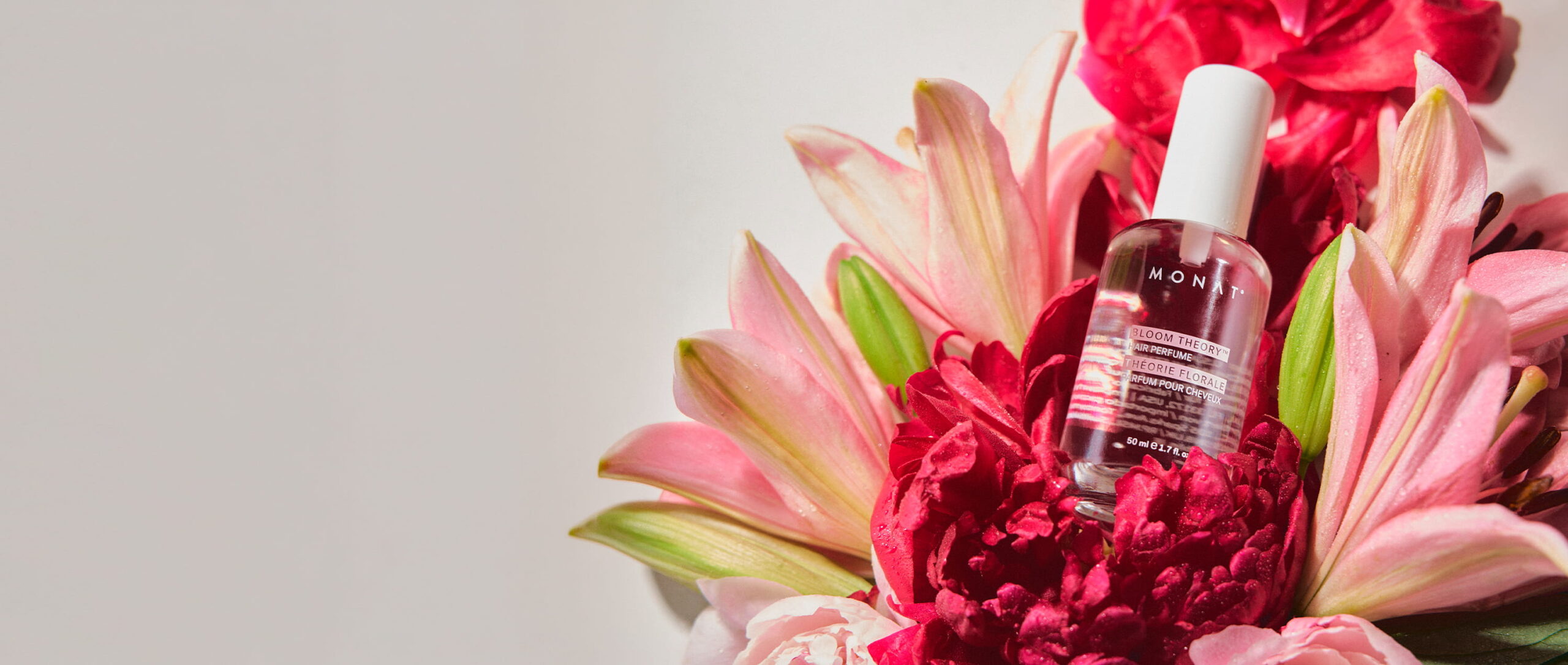 MONAT Bloom Theory Hair Perfume spray bottle nestled among pink lilies and red peonies against a light background