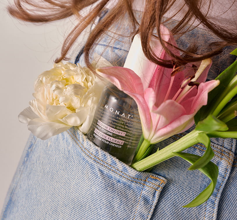 ONAT Bloom Theory Hair Perfume tucked in a denim pocket alongside a white peony and pink lily, with flowing brown hair above