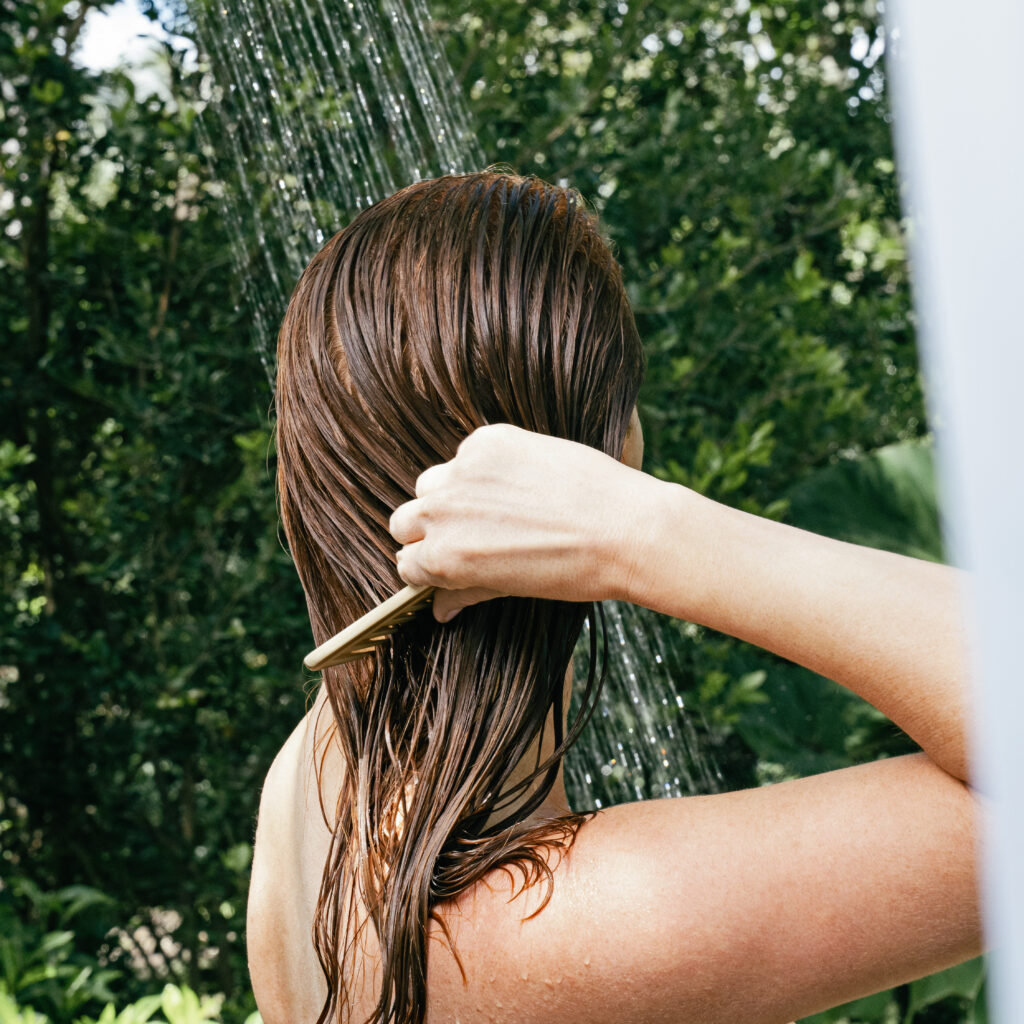 Back of a person’s head with wet brown hair being rinsed under a stream of water outdoors, with green foliage in the background
