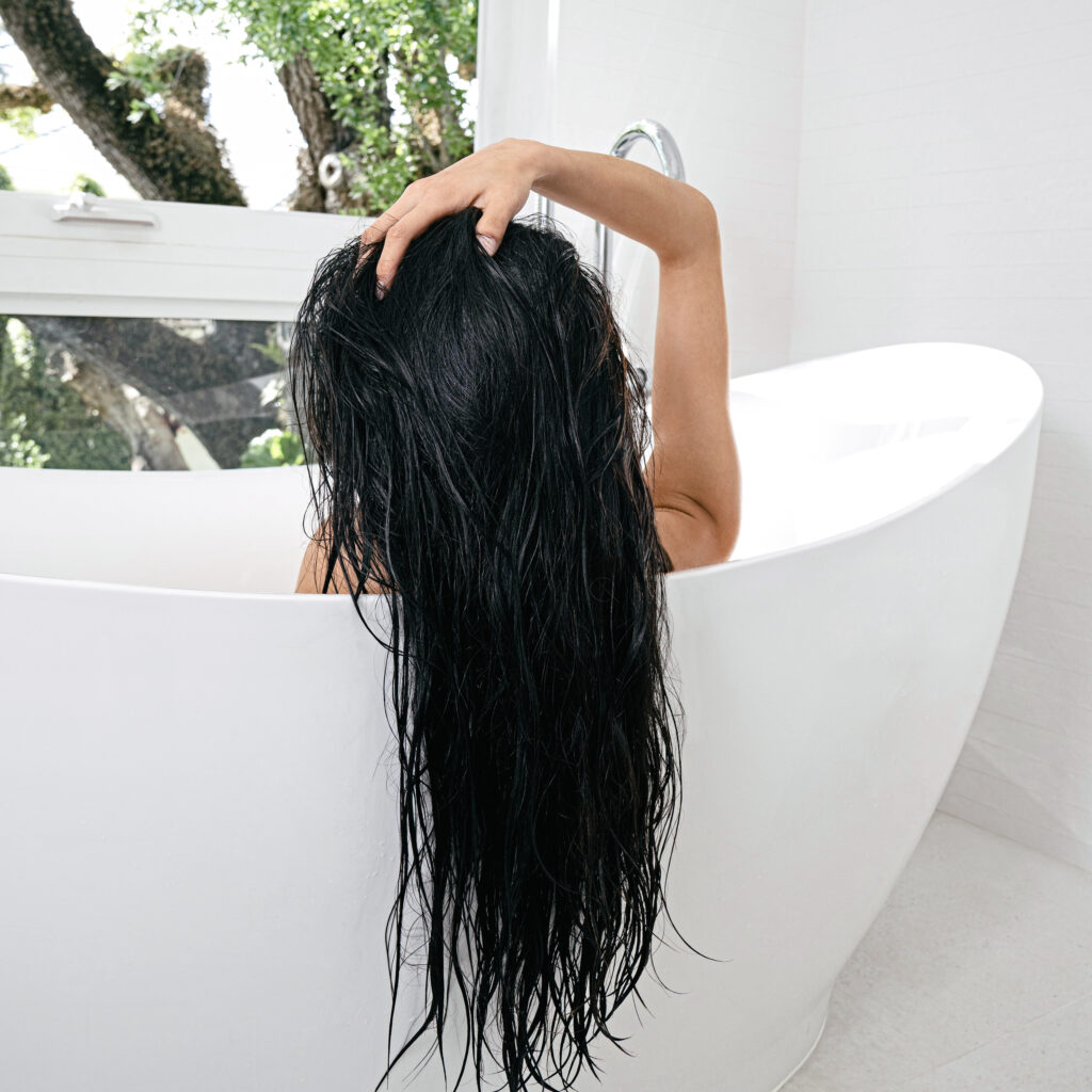 Back view of a person with long wet black hair, arm raised to hold their hair while standing in a white bathroom bathtub area. Outside greenery is visible through a window.