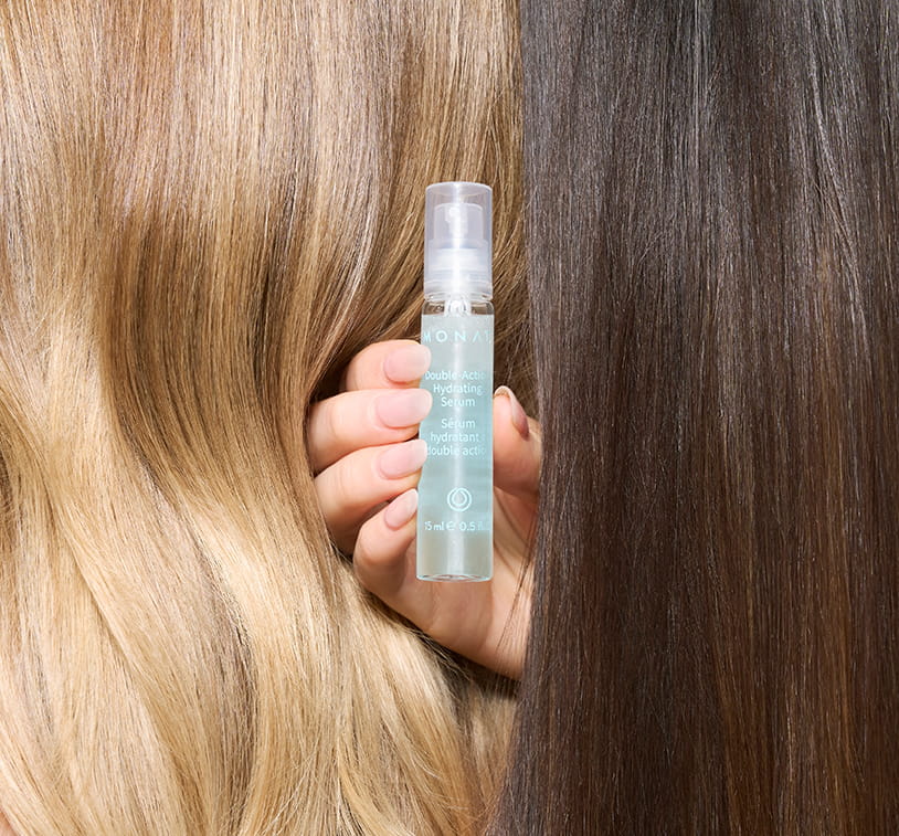 A hand holding a MONAT Double-Action Hydrating Serum vial between two contrasting hair textures — blonde wavy hair on the left and smooth dark hair on the right — highlighting the product's versatility.