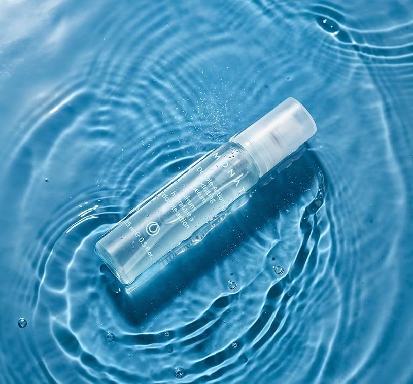 A single MONAT Double-Action Hydrating Serum vial lying on its side in blue rippling water, surrounded by concentric water rings and fine droplets, evoking deep hydration.
