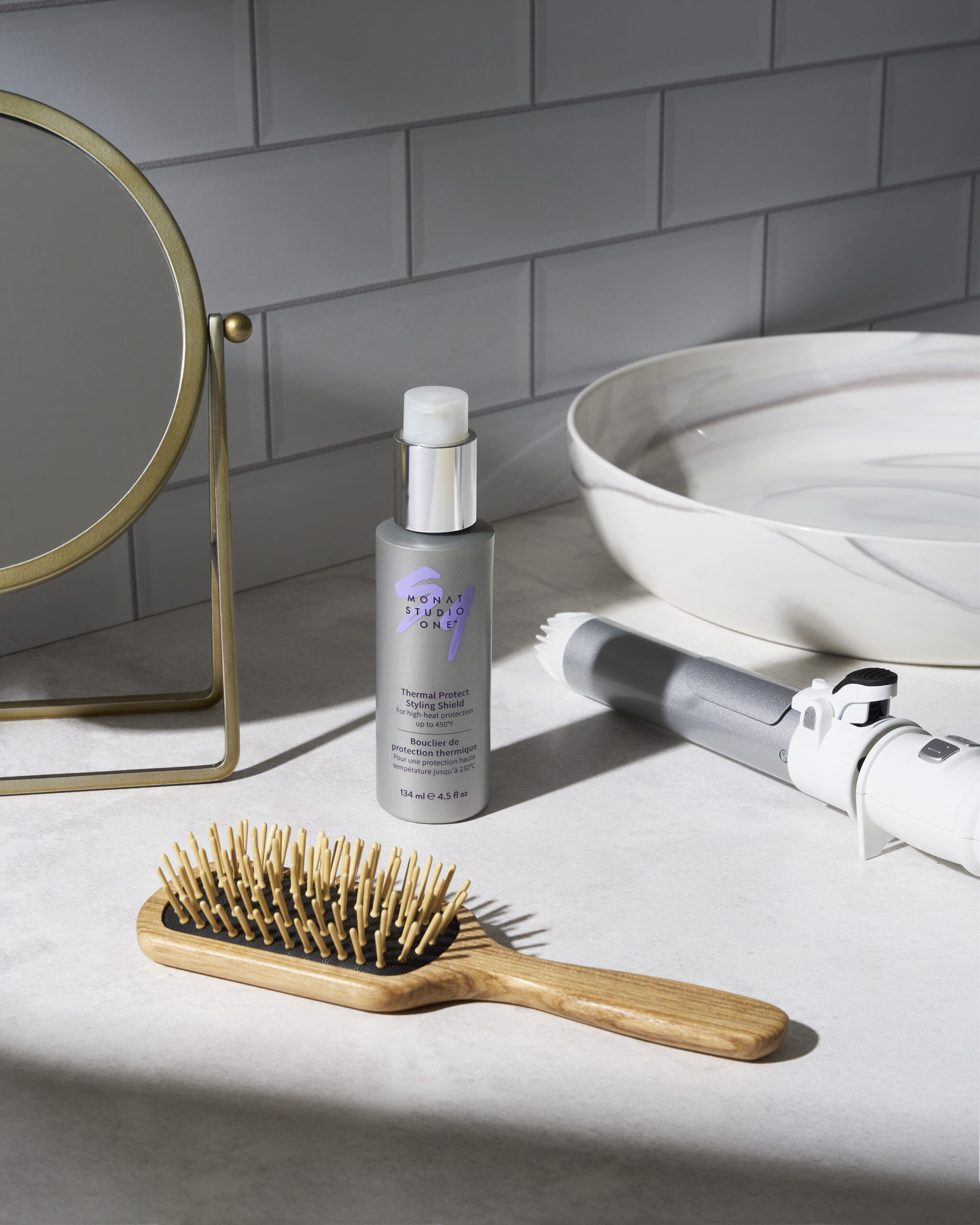 Grey tiled bathroom counter with a round gold-framed mirror, a styling product bottle, and a marble sink bowl in view. A wooden-bristle hairbrush sits in the foreground.