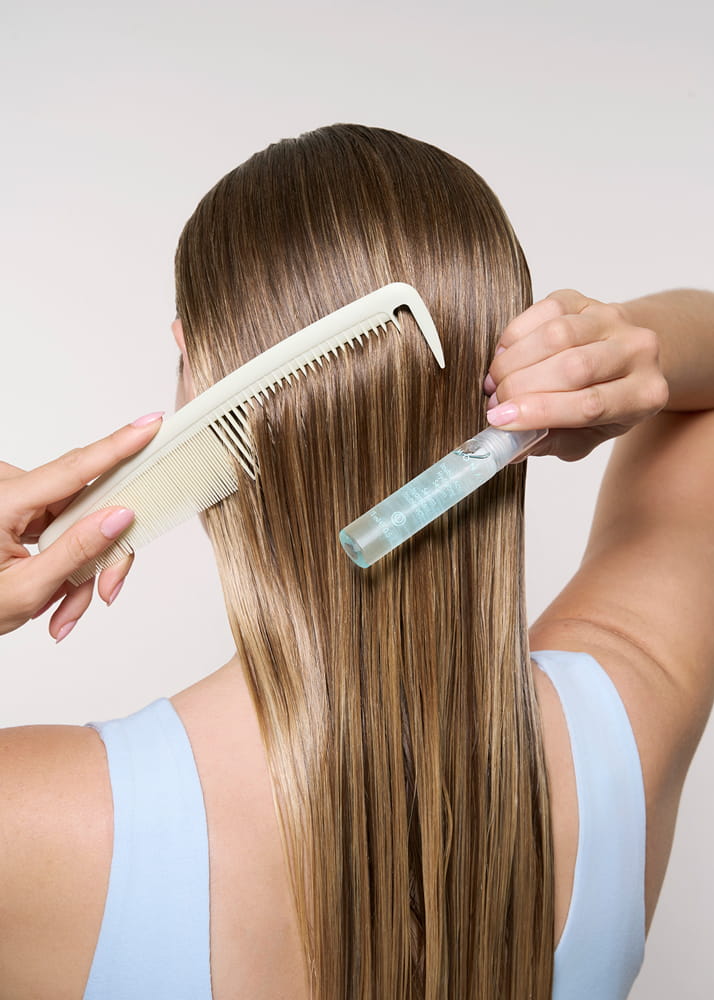 Rear view of a woman with glossy, highlighted brown hair applying MONAT Double-Action Hydrating Serum while combing it through her straight, smooth strands with a wide-tooth comb.