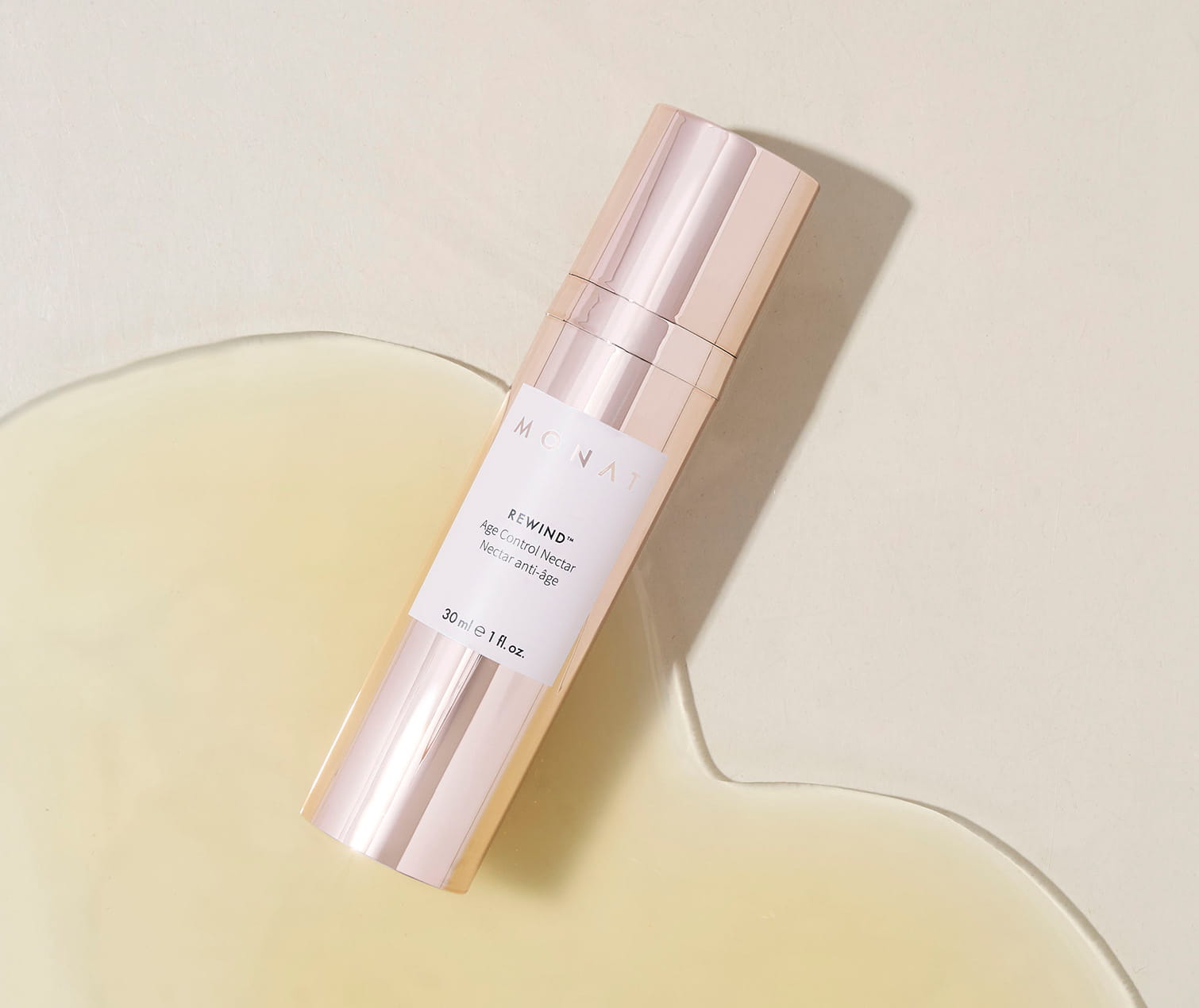 A bottle of MONAT Rewind Age Control Nectar laying flat on a neutral beige surface inside a pool of translucent golden liquid.