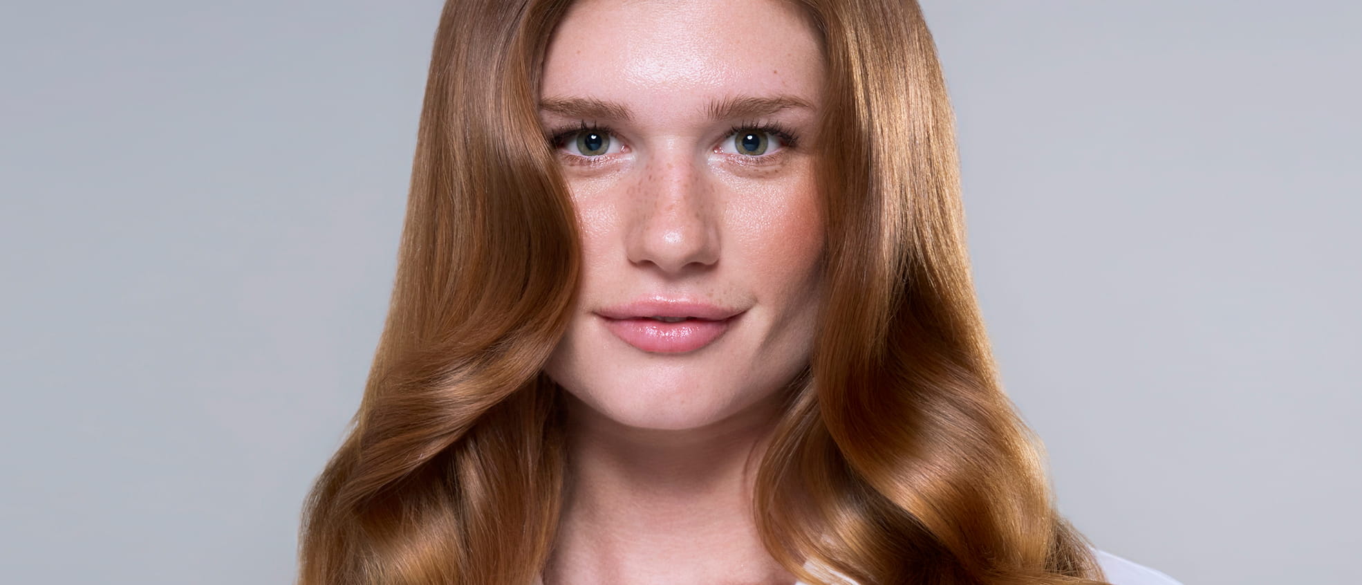 Close-up portrait of a woman with long, wavy auburn hair and freckles, looking directly at the camera against a light gray background