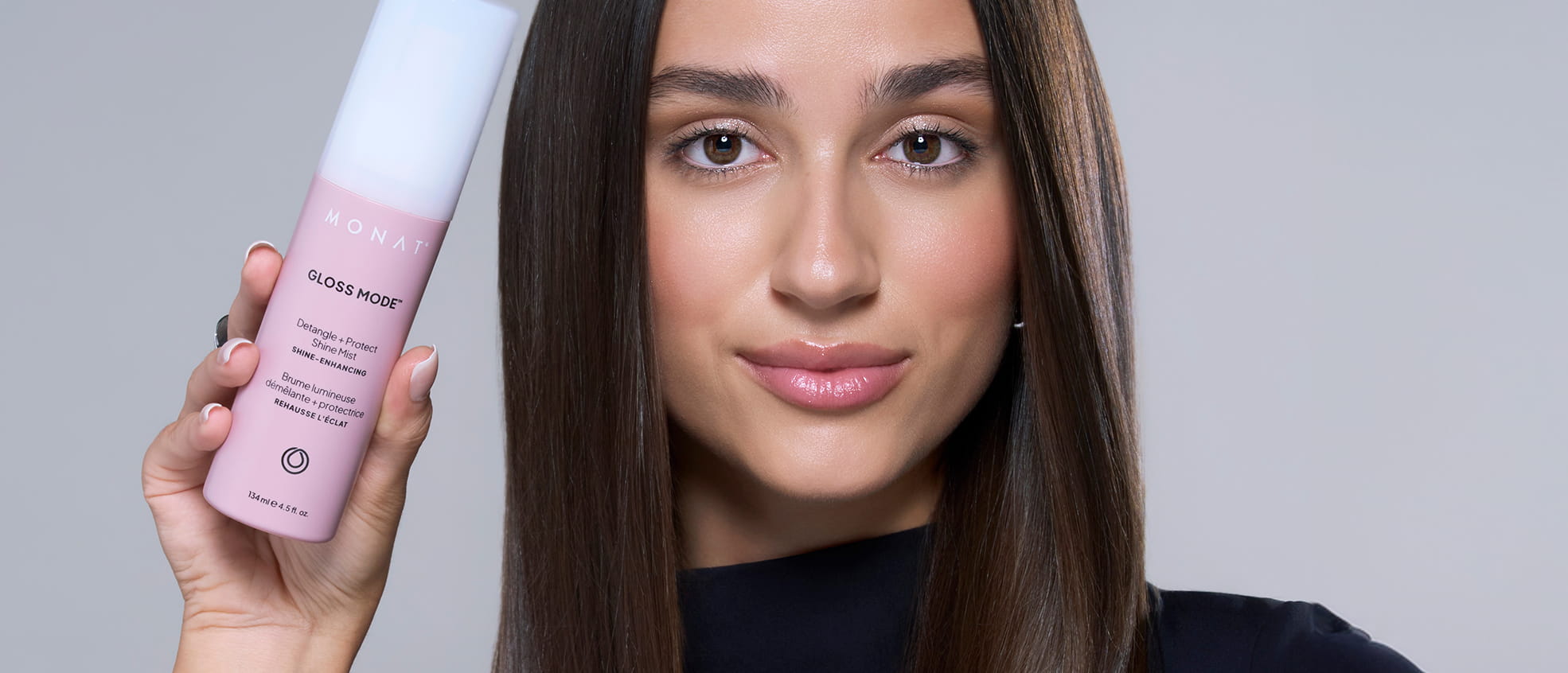 Woman holding a MONAT Gloss Mode Shine Mist bottle beside her long, straight brown hair