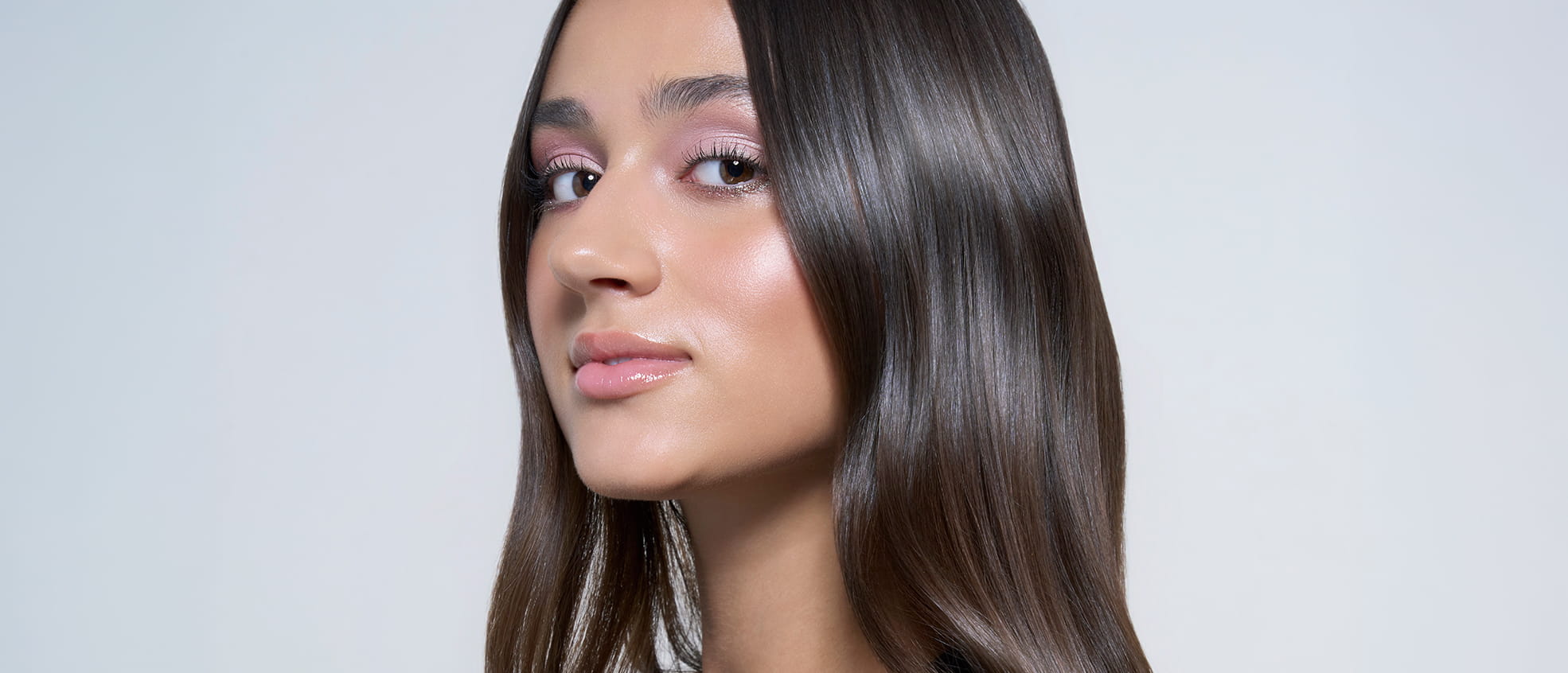 Close-up portrait of a woman with long, sleek brown hair, luminous skin, and soft pink eye makeup, highlighting a glossy, radiant finish.