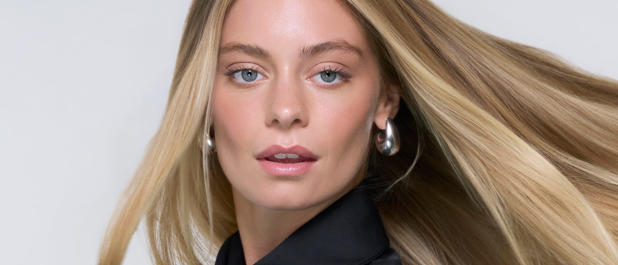 Blonde woman with long, glossy hair wearing a black top and silver hoop earrings, looking toward the camera