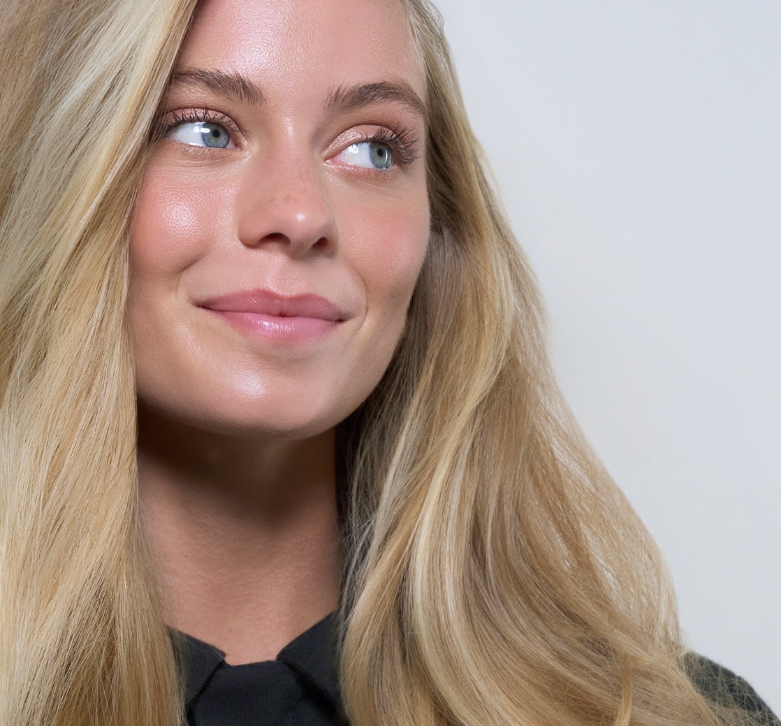 Close-up portrait of a smiling blonde woman with long, smooth, glossy hair against a light gray background.