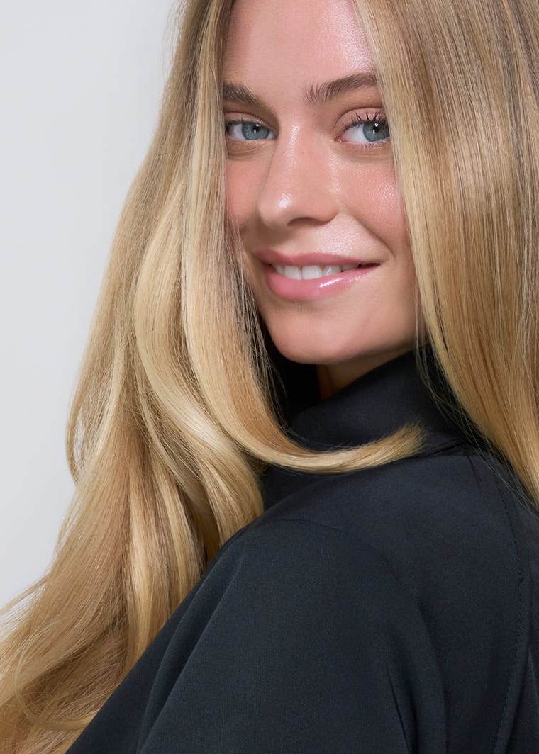 Smiling blonde woman with long, smooth hair, wearing a black top, looking over her shoulder