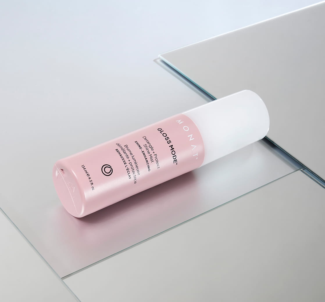 Pink MONAT Gloss Mode Detangle + Protect Shine Mist bottle lying diagonally on a reflective glass surface against a light gray background