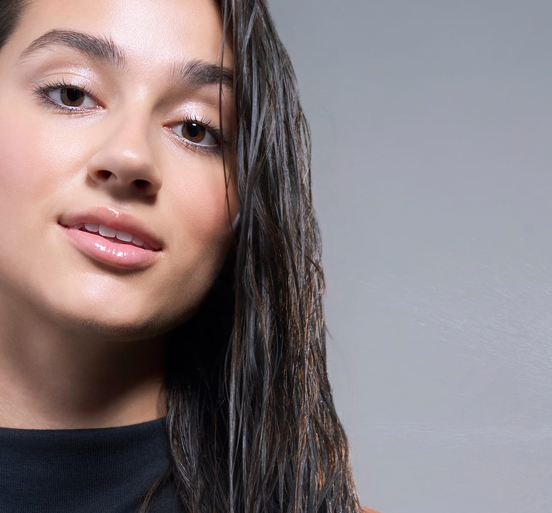 Close-up of a woman with wet, dark hair and glossy skin, with her damp hair visible along one side of her face against a soft gray background
